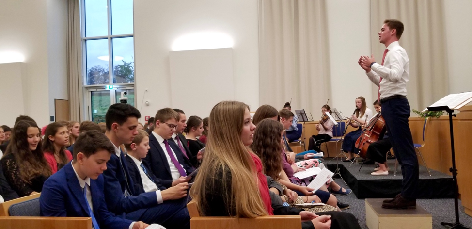 A congregação junta-se a um pequeno grupo orquestral e ao coro cantando “As Famílias Poderão Ser Eternas” no devocional para os jovens em Friedrichsdorf, Alemanha, em 19 de outubro de 2019, véspera da rededicação do Templo de Frankfurt.