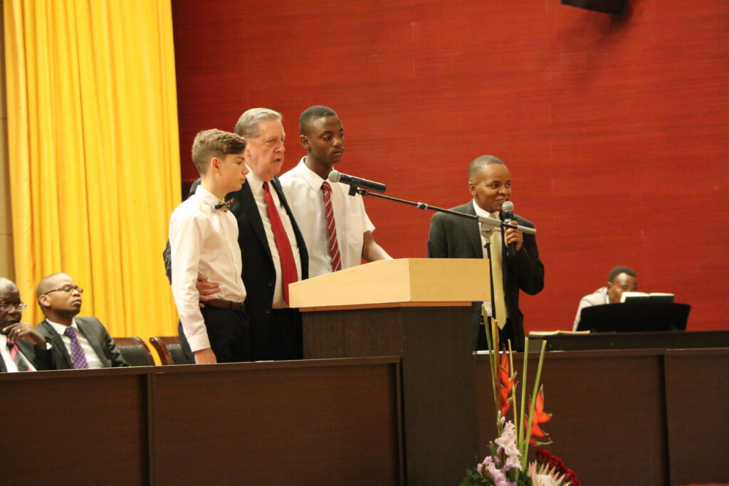 El élder Jeffrey R. Holland, del Cuórum de los Doce Apóstoles, de pie junto a dos hombres jóvenes en el púlpito mientras habla durante una conferencia multiestaca en el Centro Internacional de Conferencias Joaquim Chissano en Maputo, Mozambique, el domingo 17 de noviembre de 2019.